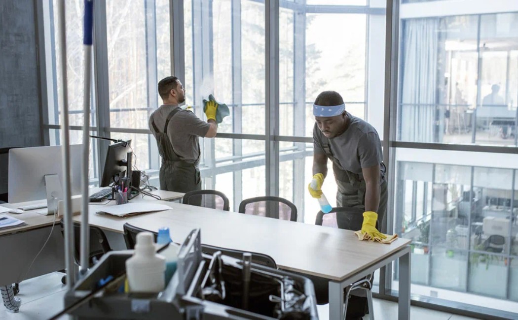 Team providing Best Cleaning Services by wiping desks and cleaning large office windows in a bright, modern workspace.