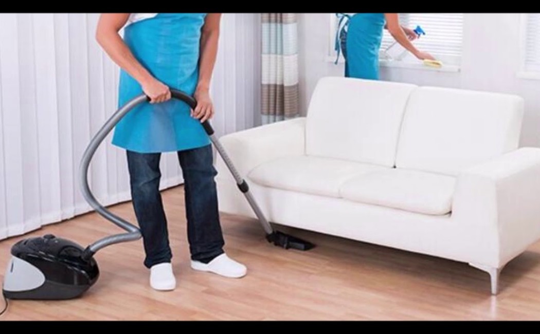 Professional cleaners vacuuming a living room floor while another team member dusts surfaces near a white sofa, delivering the best cleaning services.