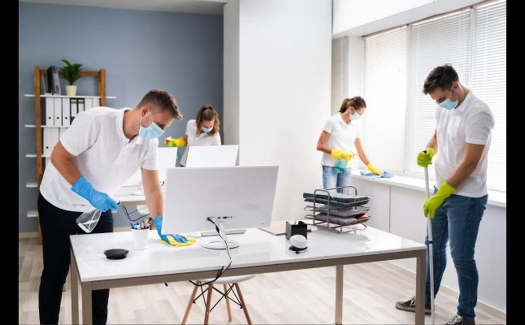 Team providing Office Cleaning Services disinfecting desks, windows, and floors in a modern workspace while wearing gloves and masks.