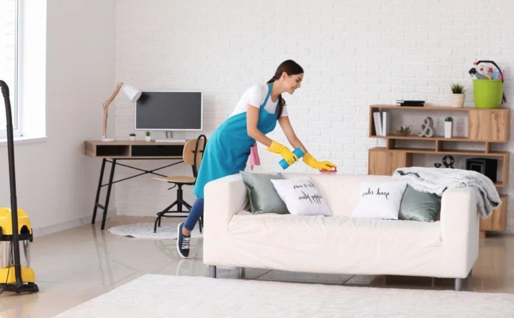 Apartment cleaning professional dusting a white sofa in a bright and modern living room.