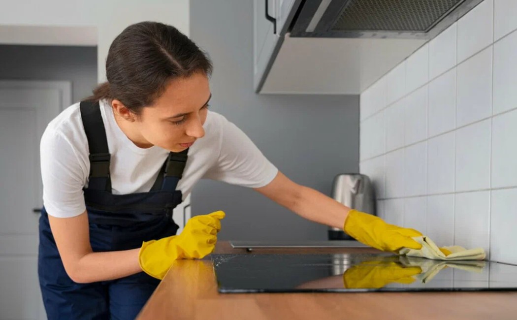 A cleaner wearing gloves wipes the stovetop and backsplash during professional Kitchen Cleaning Services in a modern kitchen.