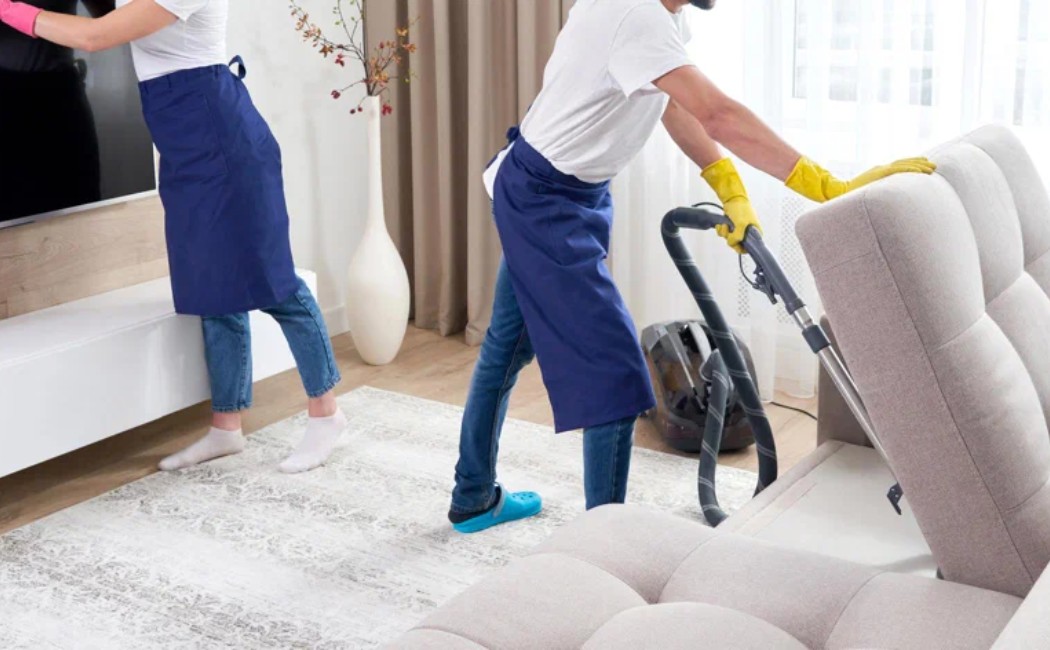 Professional apartment cleaning services team vacuuming and dusting a modern living room sofa and entertainment area.
