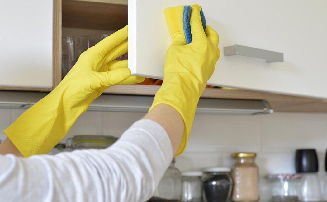 Person wearing yellow gloves wiping a kitchen cabinet door with a sponge, representing professional Kitchen Cleaning Services.