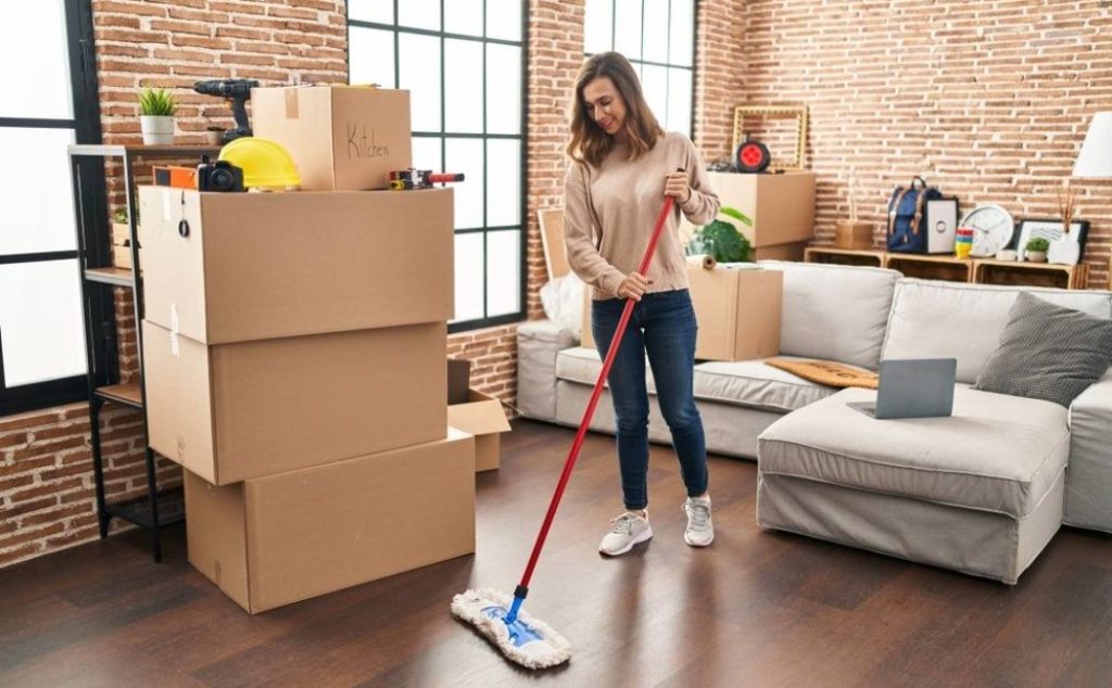 Move-Out Cleaning service in progress as a woman mops the floor of a living room surrounded by packed moving boxes and household items.
