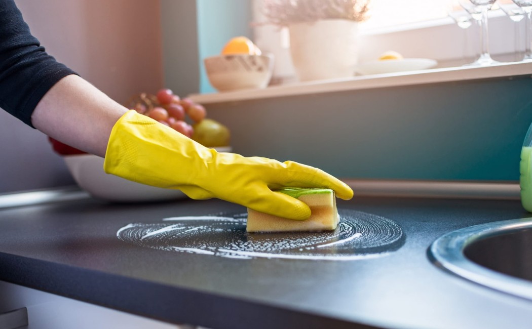 Person wearing yellow gloves performing kitchen cleaning by scrubbing a modern kitchen countertop with a sponge and cleaning solution.
