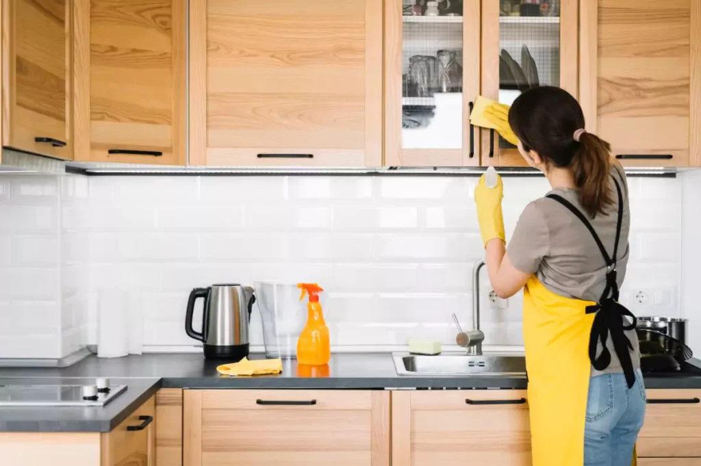 Woman in yellow apron and gloves cleaning kitchen cabinets and sink during a thorough Kitchen Cleaning routine.